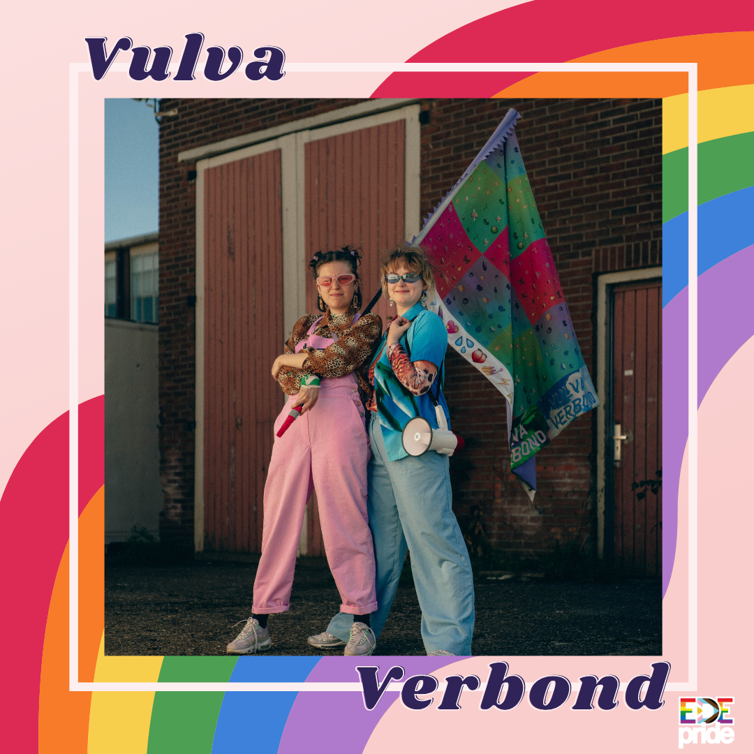 Afbeeldingsbeschrijving voor toegankelijkheid: Foto van twee vrouwen die voor een stenen muur staan. De linker vrouw draagt een roze tuinbroek en een shirt met tijgerprint. Ze heeft haar armen over elkaar en glimlacht in de camera. Ze draagt een roze zonnebril en heeft kort donker haar met knotjes. De rechter vrouw draagt een blauwe broek en blauwe blouse met print. Ze draagt een blauwe zonnebril en glimlacht in de camera. Ze heeft kort donkerblond haar. Achter de vrouwen is een groen, paars, roze geblokte vlag te zien. In paarse letters staat bij de foto Vulva Verbond.