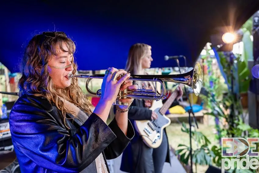 Een foto van twee vrouwen die op een podium optreden. De linker vrouw heeft halflang krullend lichtbruin haar en speelt met haar ogen dicht op een trompet. Ze draagt een zwart-leren jas en draagt een gezicht-sieraad over haar neus en wangen. De andere vrouw heeft een lange zwarte jas aan, heeft halflang blond haar en speelt op een elektrische gitaar.